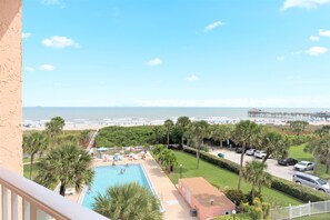 A pool with a view of the beach and pier, surrounded by palm trees and a parking area. - Stylish Coastal Penthouse | Oceanfront Views  | Heated Pool  | Steps to the Pier (Cocoa Beach)