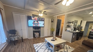 A living room with a television, a sofa, a coffee table, and a kitchen area in the background.