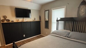 A bedroom with a bed, a ceiling fan, a TV mounted on the wall, a window with blinds, and a wooden headboard.