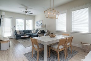 A modern living room with a dining area, a ceiling fan, and large windows with blinds.