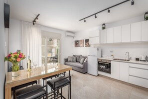 A modern kitchen with white cabinets, a dining area with a glass table, and a gray sofa. - Stylish one BDR with balcony (Jerusalem)