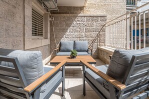 A sunlit outdoor seating area with a stone wall backdrop, a wooden table, and grey upholstered chairs.