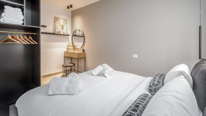 A hotel room with a neatly made bed, a wardrobe with hanging clothes, a small desk with a chair, and a wall-mounted mirror. - Stylish one BDR with balcony (Jerusalem)
