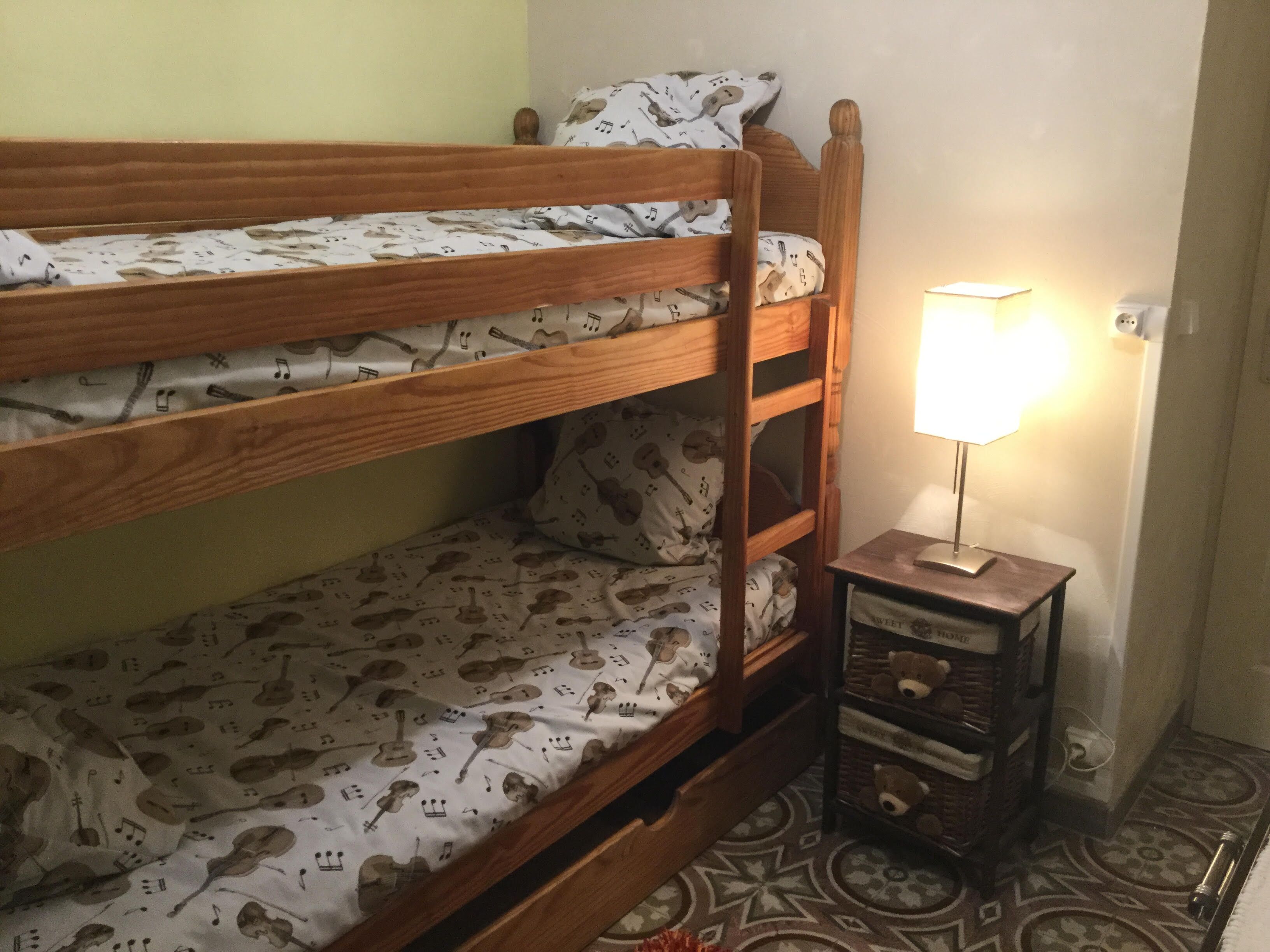 A bunk bed with a patterned sheet, a bedside table with a lamp, and a patterned floor.