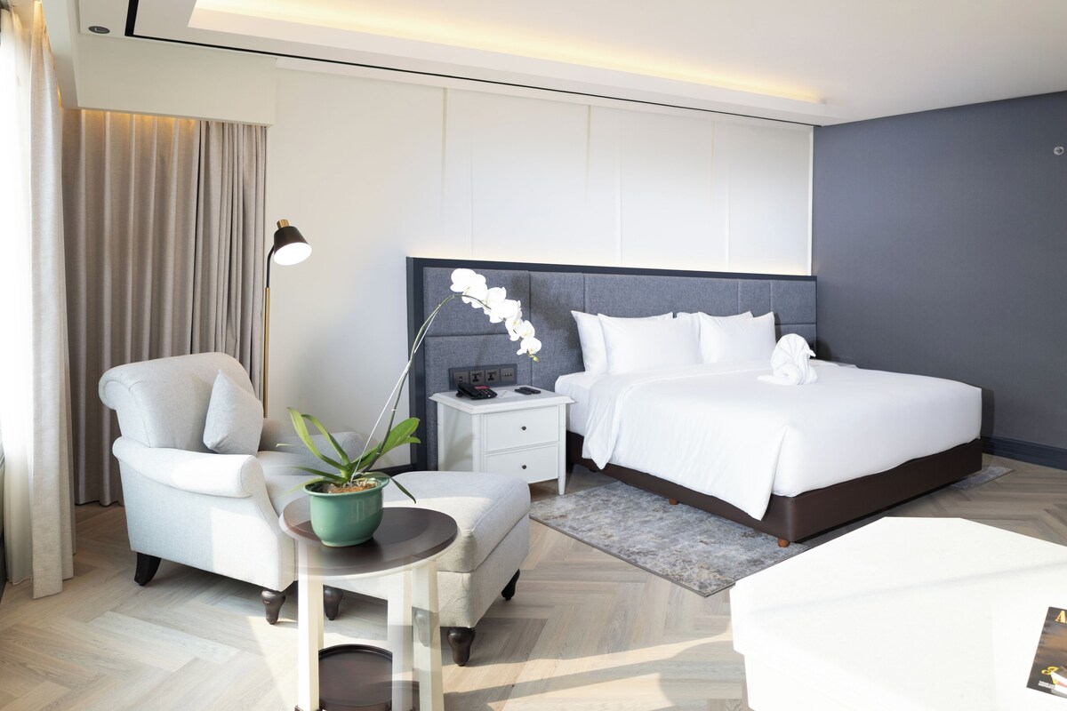 Habitación de hotel moderna con cama, mesita de noche, lámpara, sillón y planta en maceta.