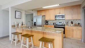 Une cuisine moderne dotée de plans de travail en granit, d’armoires en bois, d’un réfrigérateur en acier inoxydable, d’un four à micro-ondes, d’une cuisinière et de trois tabourets de bar.
