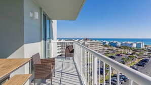 Un balcon avec des fauteuils en osier et une table en bois, d’où l’on peut admirer un parking et les bâtiments situés en bord de mer.