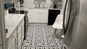 Una cuina amb armaris blancs, un paviment amb dibuixos en blanc i negre, un frigorífic d'acer inoxidable i un rentapeus amb un frontal de taulells decoratius.