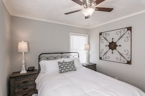 Quarto com cama, duas mesas de cabeceira, ventoinha no teto, janela com persianas e um objeto decorativo na parede.