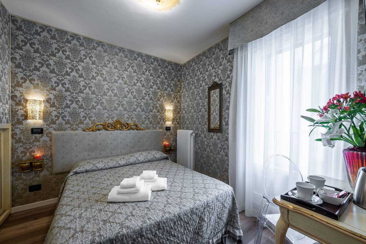A hotel room with a patterned wallpaper, a bed with neatly folded towels, a bedside table with cups and a tray, and a vase with flowers.