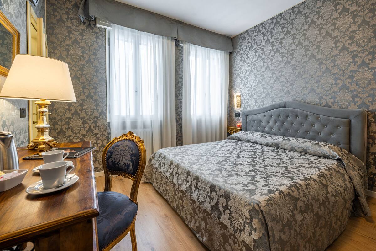 A hotel room with a large bed, a desk with a lamp, a chair, and a window with curtains.