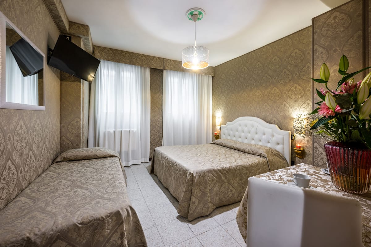 A hotel room with two beds, a television mounted on the wall, a chandelier, and a table with a vase of flowers.