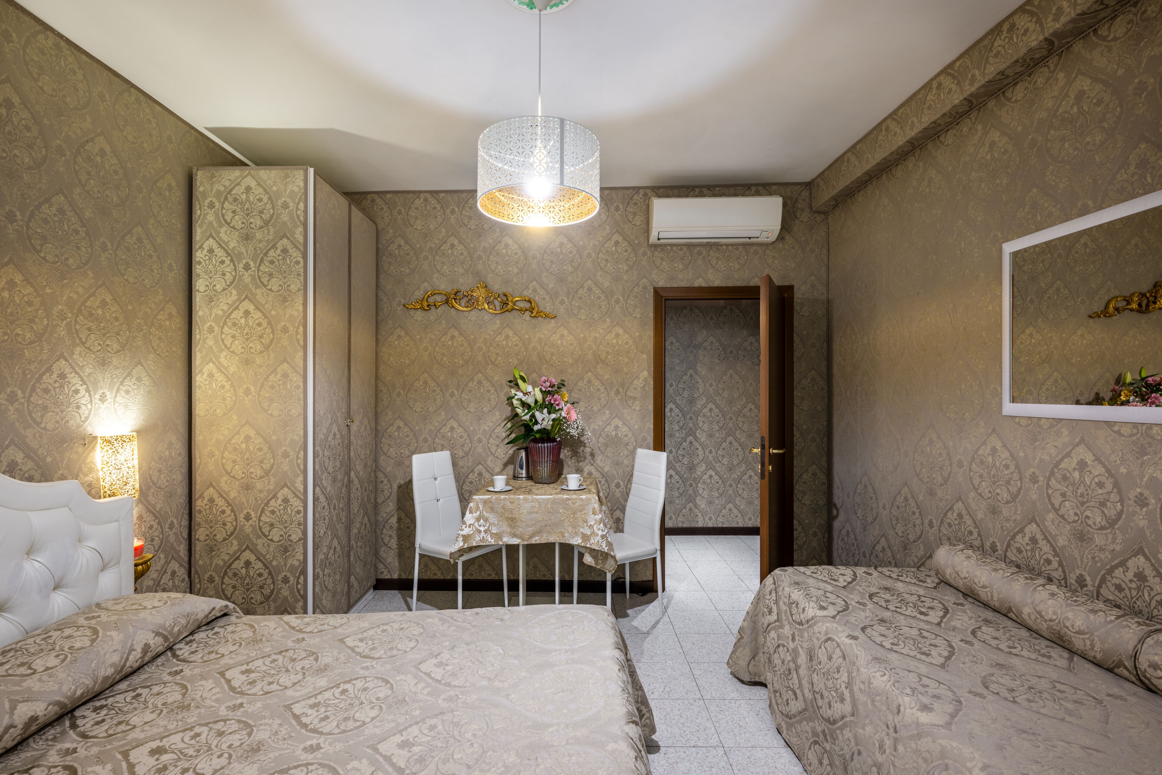 A hotel room with a bed, a small dining table with chairs, a decorative wall hanging, and an air conditioning unit.