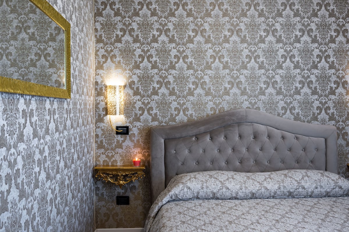 A bedroom with a tufted headboard, patterned wallpaper, a bedside table with a candle, and a framed mirror.