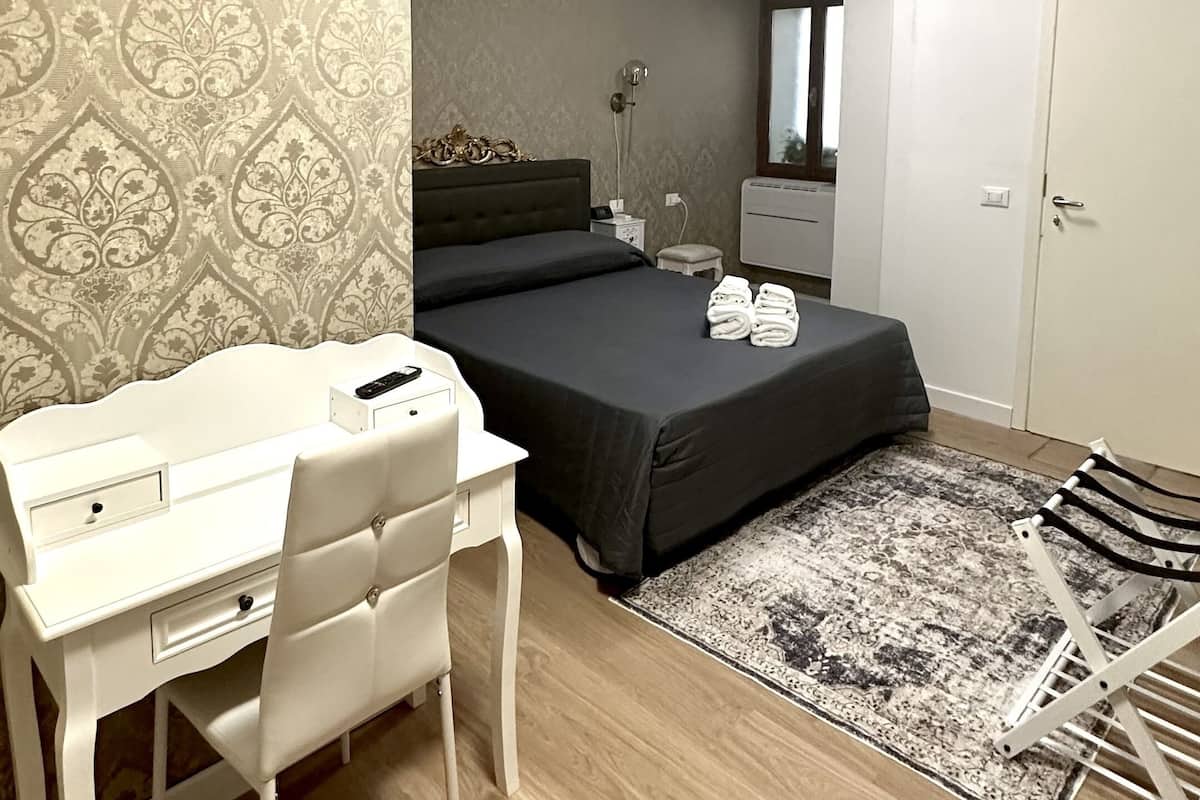 A bedroom with a bed, a desk, and a chair. There is a window, a radiator, and a door.