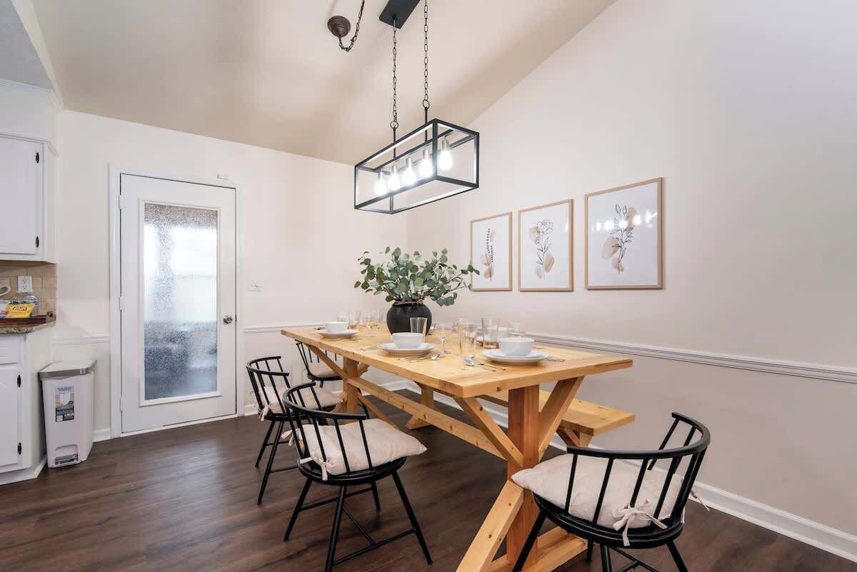 A dining area with a wooden table set for four, black chairs with cushioned seats, a hanging light fixture, and framed botanical prints on the wall.