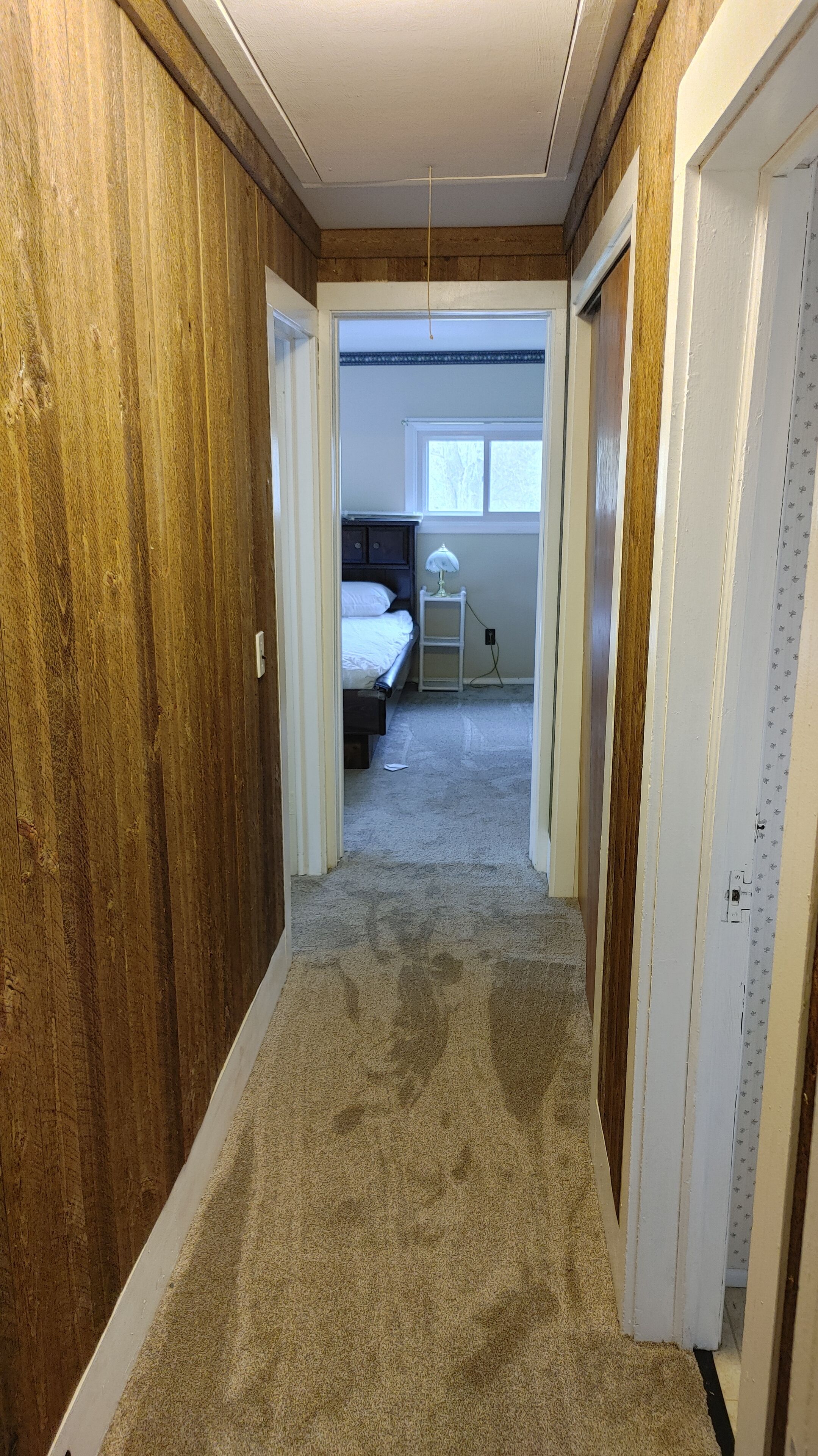 A narrow hallway with wooden paneling on the walls, carpeted floor, and a bedroom with a bed, nightstand, and window in the background.