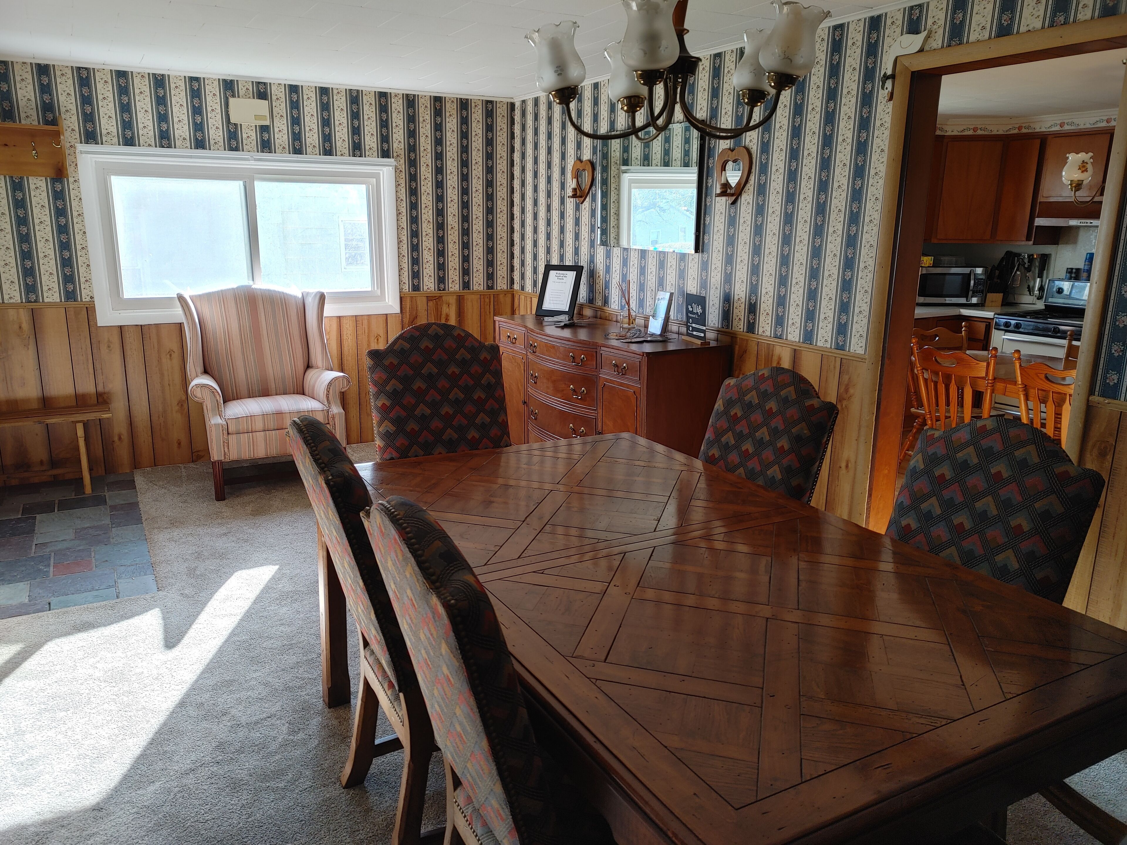 A dining area with a wooden table and chairs, a chandelier, and a kitchen visible through an open doorway.