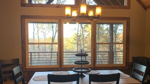 Un coin repas avec une table et des chaises en bois, de grandes fenêtres donnant sur la verdure et un lustre.