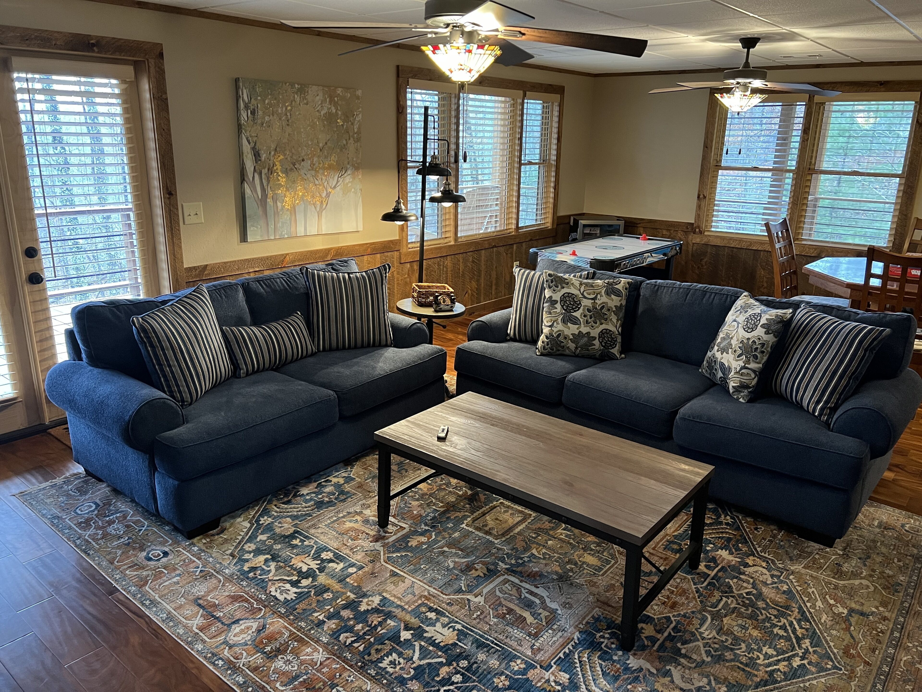 A living room with a sectional sofa, a coffee table, and a rug. There are two windows with blinds, a ceiling fan, and a painting on the wall.