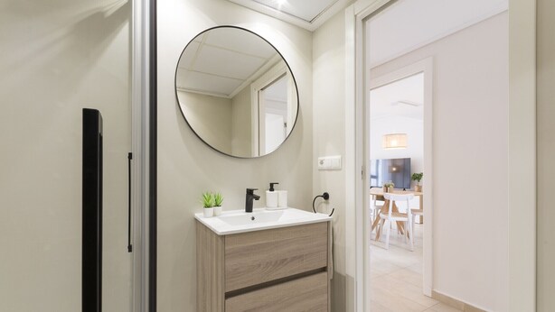 Un baño moderno con un espejo redondo, un tocador de madera y vistas al comedor contiguo.