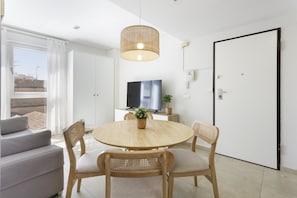 Un salon moderne avec une table ronde en bois, quatre chaises en bois, un canapé, un téléviseur et une grande fenêtre avec des rideaux.