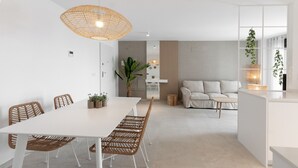 A modern, minimalist dining area with a white table and wicker chairs, a hanging light fixture, and a view into a living space with a sofa and plants. - Amazing holidays in this  New condo mediterranean style. Wifi - only families (Playa de Gandía)