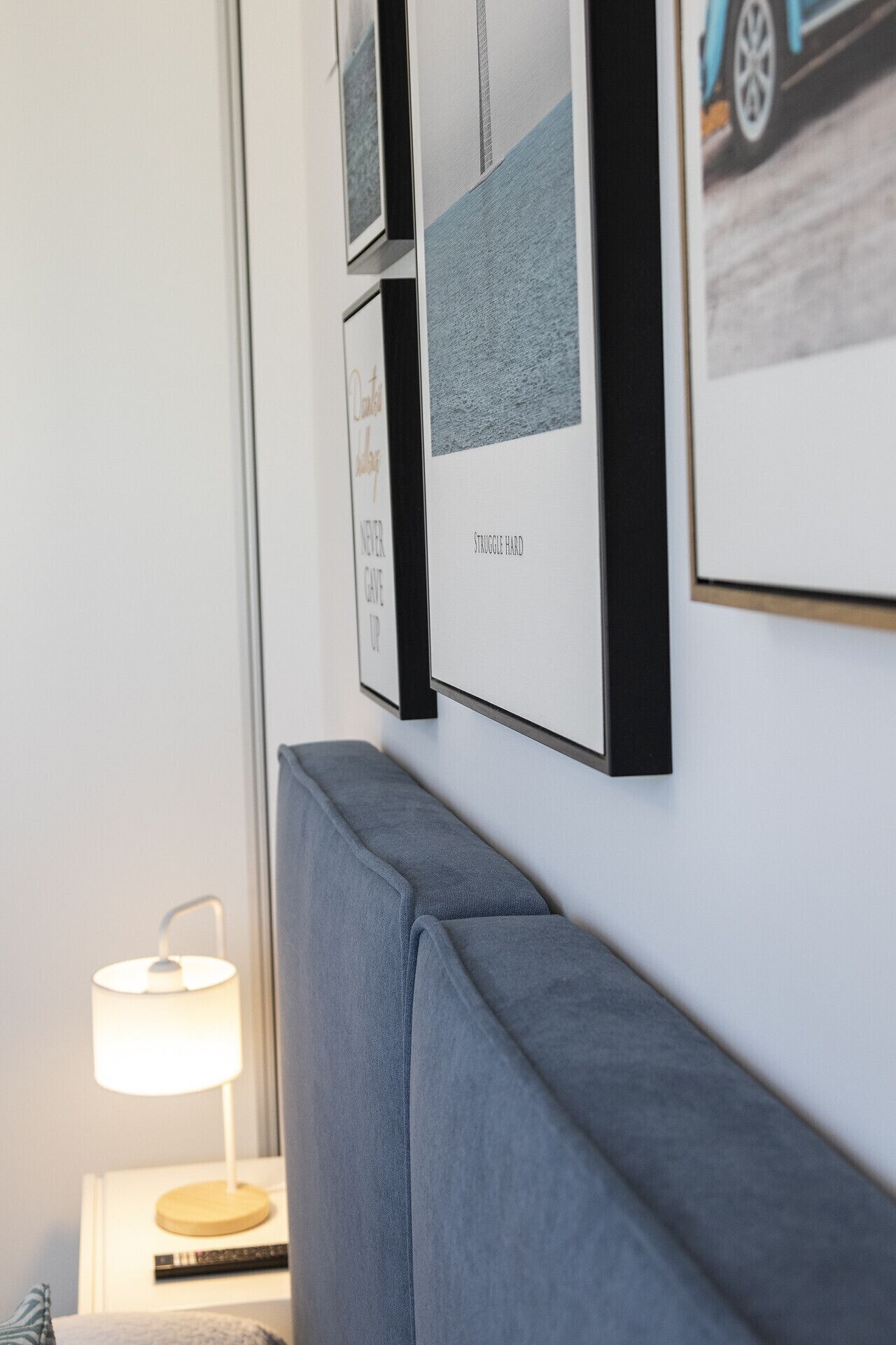 Quarto moderno com cabeceira estofada em cinza, mesa de cabeceira com abajur e quadros nas paredes.