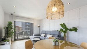 A modern living room with a glass dining table, grey sofa, and a large window offering a city view.