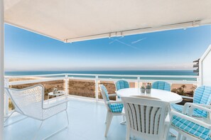 A balcony with a white table and chairs, overlooking a beach and ocean.