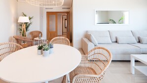 Una sala de estar moderna con una mesa redonda blanca, sillas de mimbre, un sofá de tonos claros y un ventanal con vistas a la vegetación.