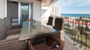 Un balcón con mobiliario de mimbre, una mesa de vidrio y vistas al océano y edificios.