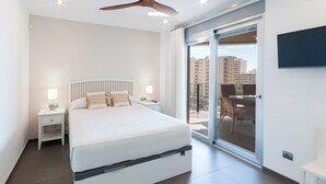 Un dormitorio moderno con una cama grande, una mesita de noche, un ventilador de techo y un puerta corrediza de vidrio que da a un balcón exterior.