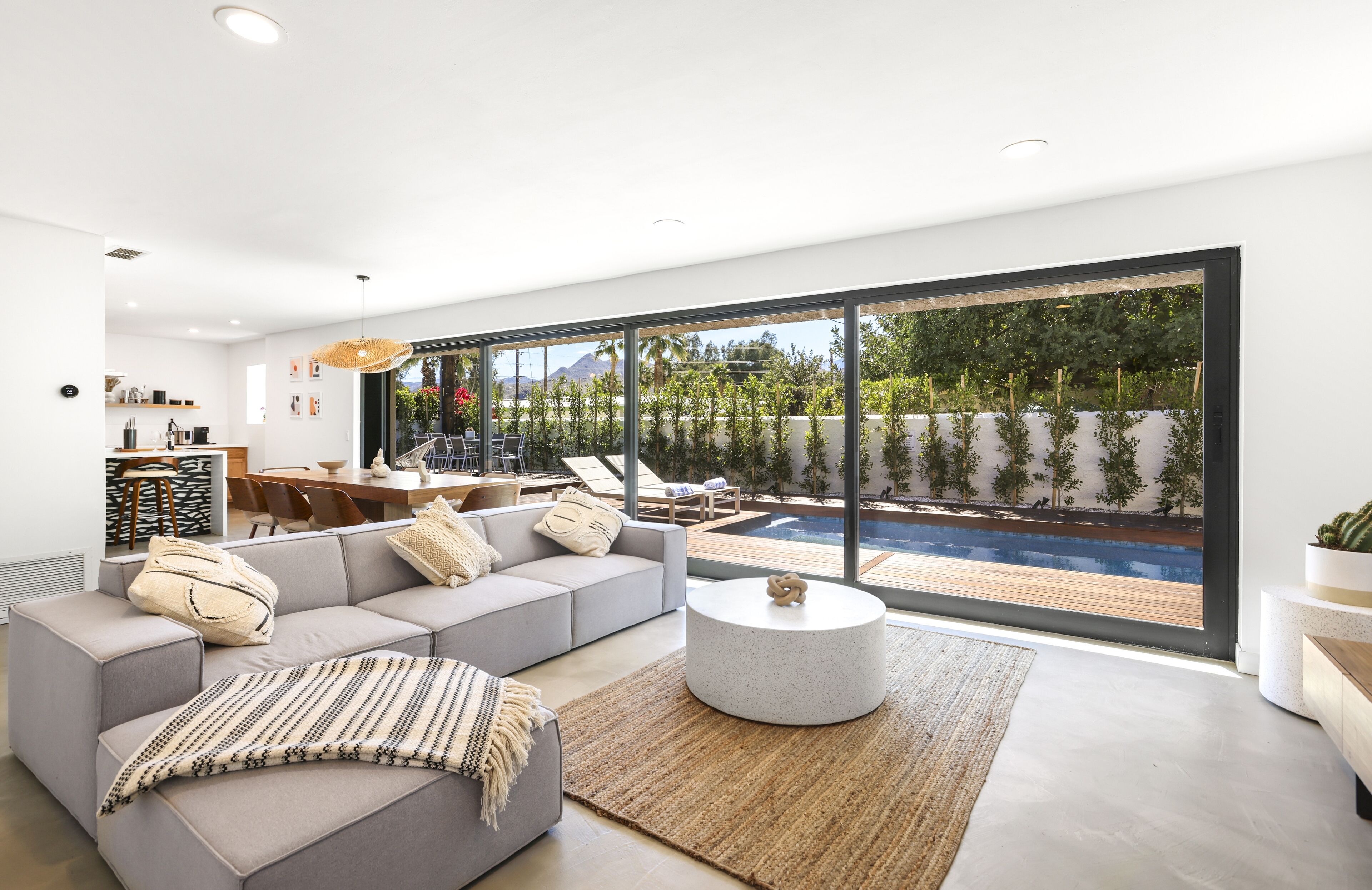 A modern living room with a large window, a gray sofa, a round coffee table, and a dining area with a pool view.