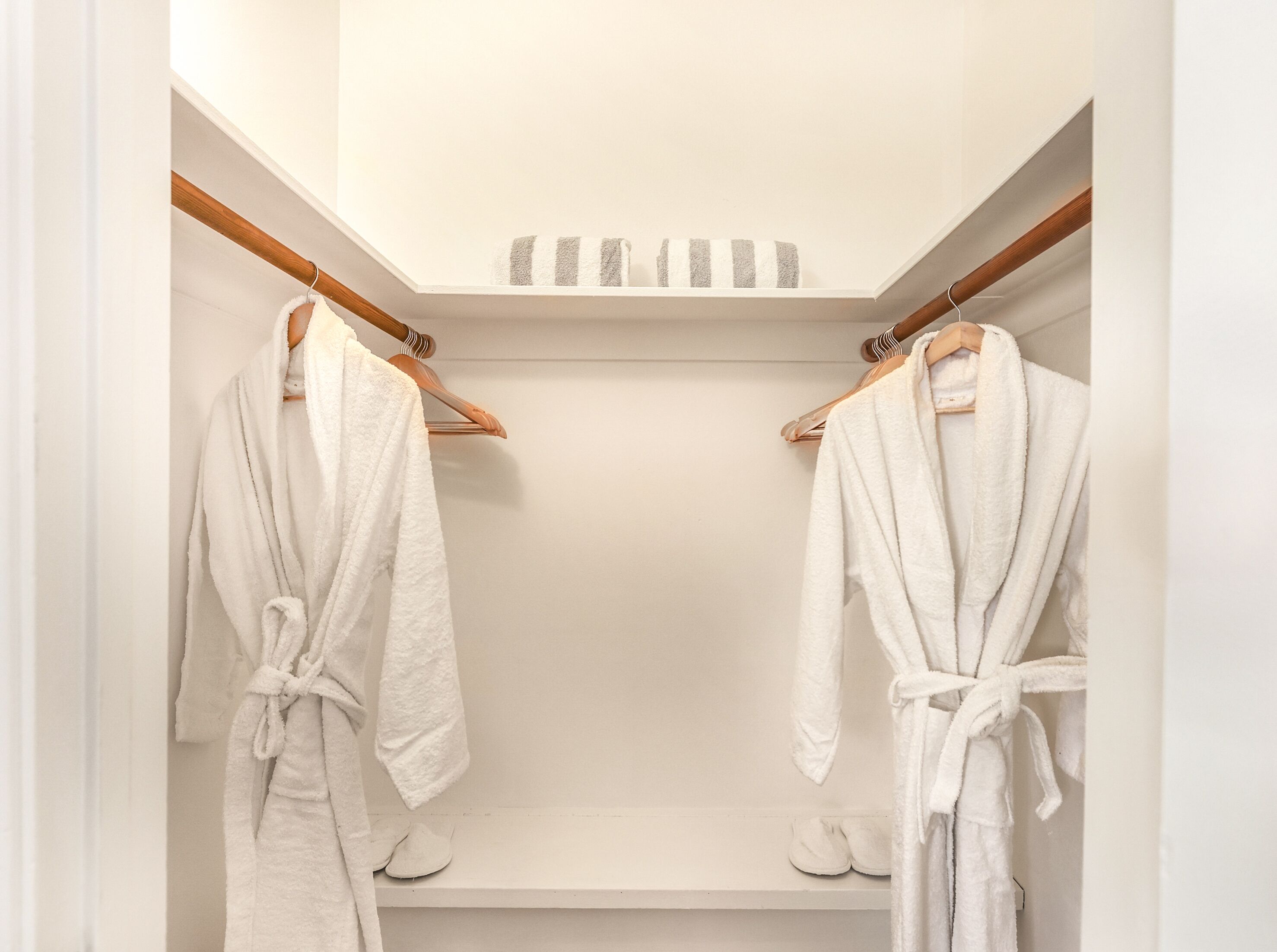 A walk-in closet with two white robes hanging on wooden racks, a wooden ceiling, and a white wall.