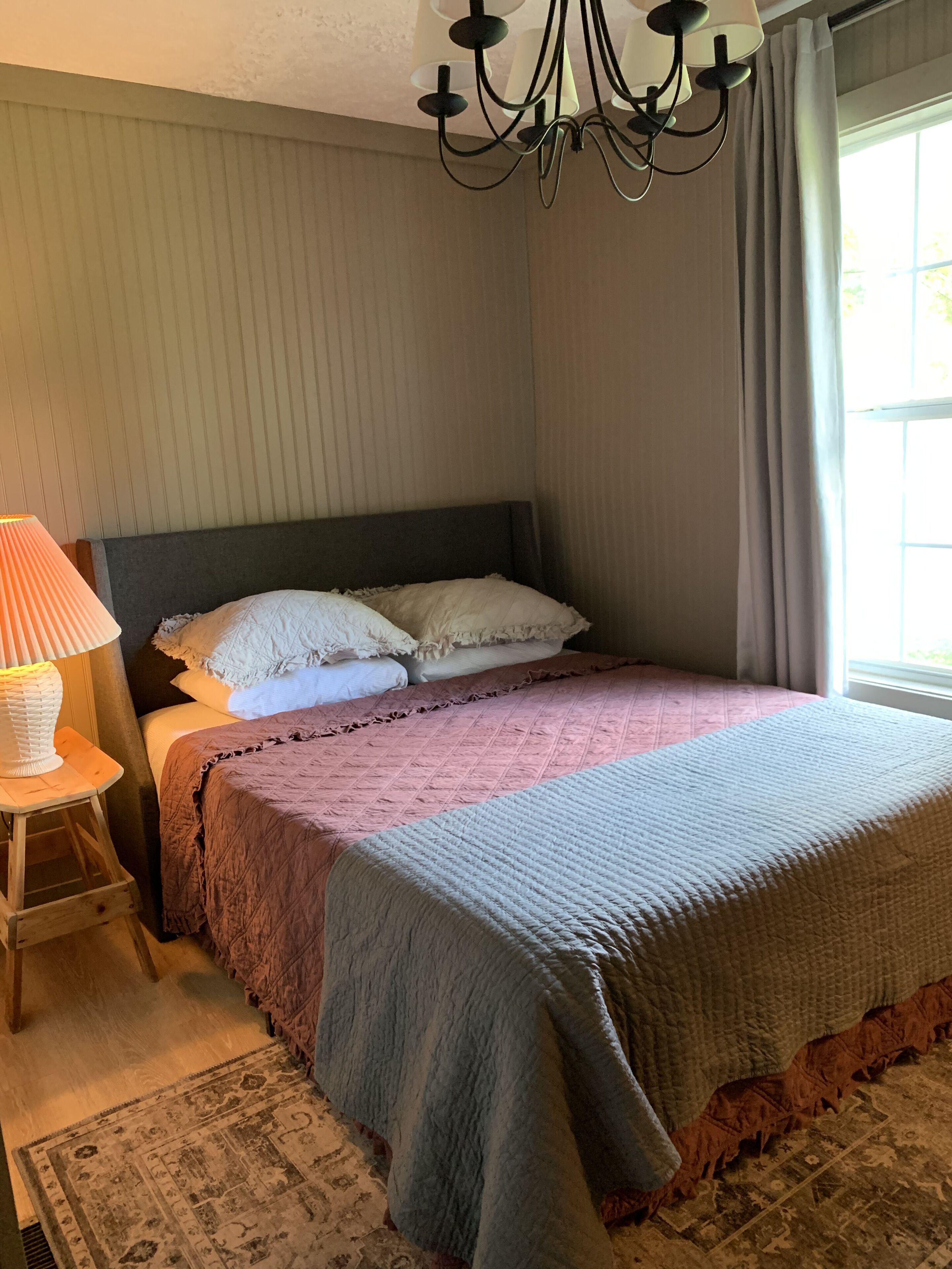 A bedroom with a bed, a nightstand, a lamp, a chandelier, and a window with curtains.