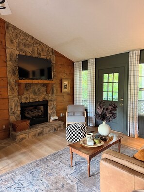 A living room with a stone fireplace, a television mounted on the wall, a checkered ottoman, a wooden coffee table, and a view of greenery outside.