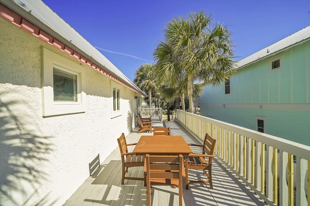 A covered outdoor area with a wooden table and chairs, a white railing, and palm trees in the background.