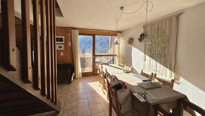 A cozy room with a wooden staircase, a dining table with chairs, a view of mountains through a large window, and a ceiling fan. - Spacious, comfortable apartment at the foot of the slopes (Arvieux)