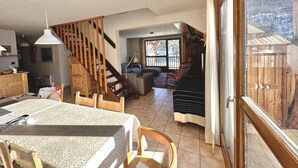 A spacious living area with a wooden staircase, a dining table, and a view of a snowy landscape through a large window. - Spacious, comfortable apartment at the foot of the slopes (Arvieux)