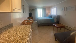 A compact room with a bed, a small table, a chair, and a kitchenette area.