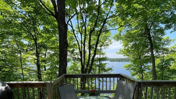 A wooden deck with a glass table and four chairs, overlooking a lake with trees in the background. - Steps from Beautiful Lake Michigamme (Michigamme Township)