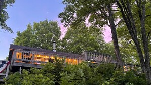 A rustic building with a wooden deck and a chimney, surrounded by lush greenery. - Steps from Beautiful Lake Michigamme (Michigamme Township)