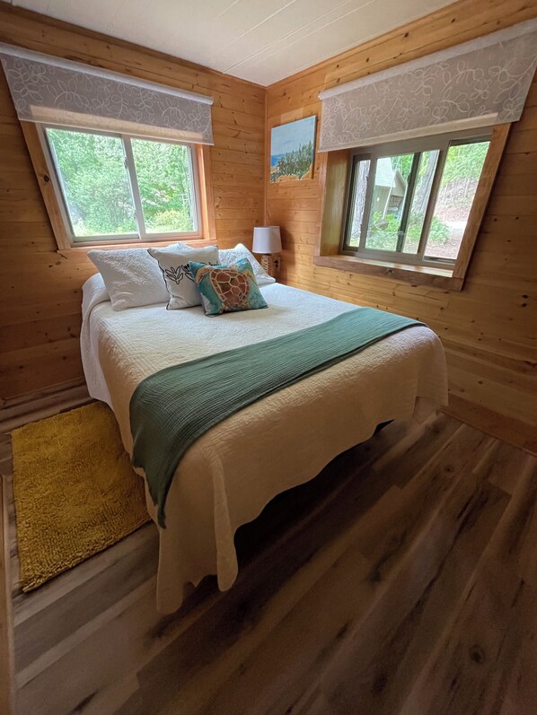 A wooden cabin bedroom with a bed, two windows, a nightstand, and a lamp.