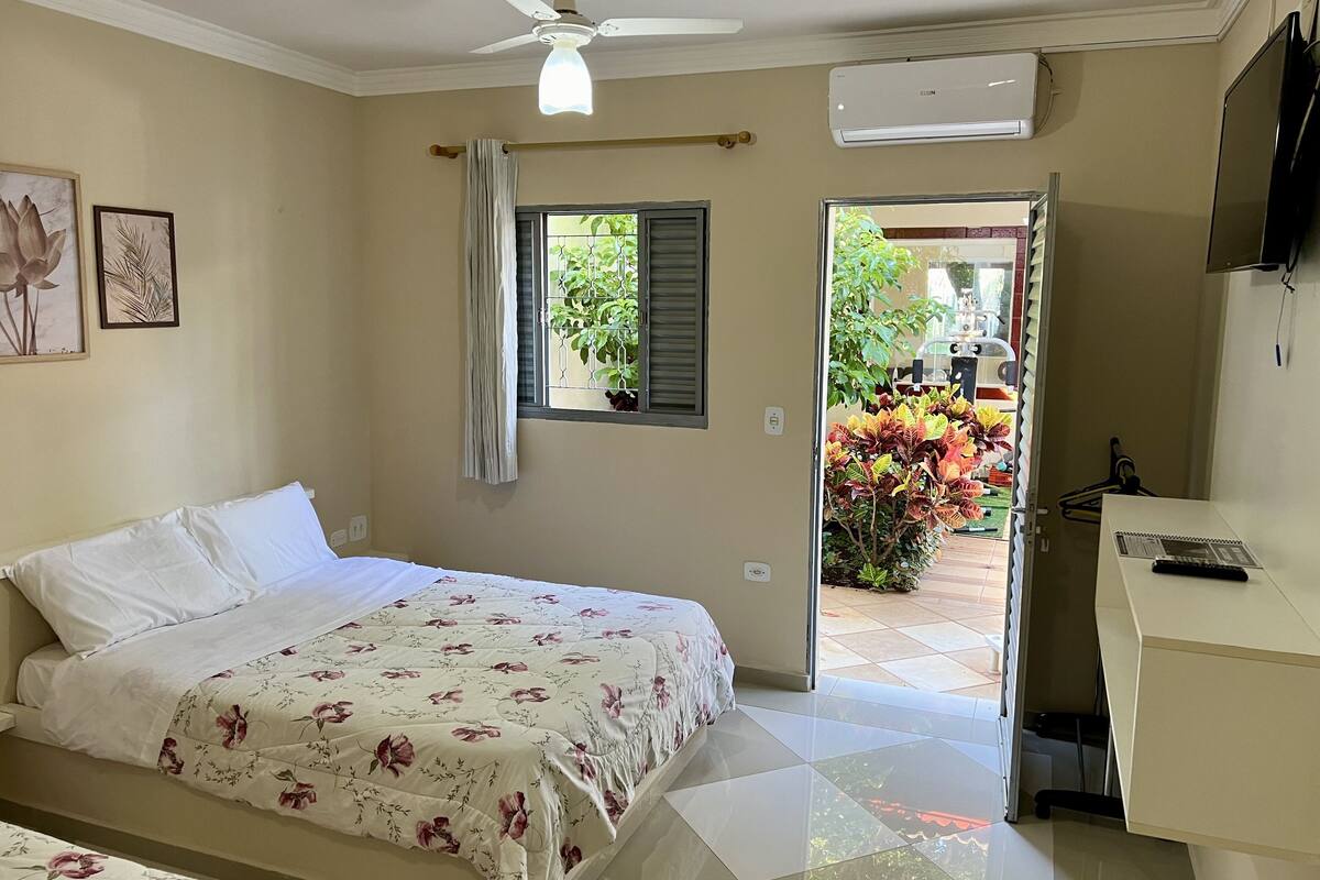A bedroom with a bed, a ceiling fan, an air conditioner, a window with curtains, and a view of a garden outside.