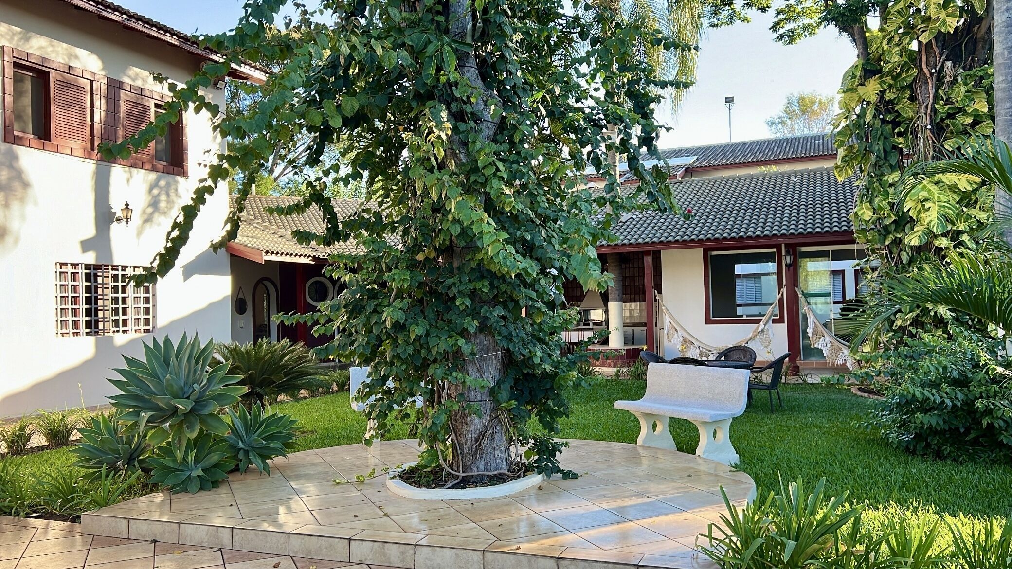 Uma casa com telhado de cerâmica, um jardim com um banco e uma árvore grande.