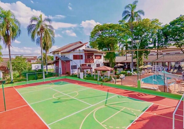A sports court with a basketball hoop, a pool area with lounge chairs, and a building with a red roof and white walls.