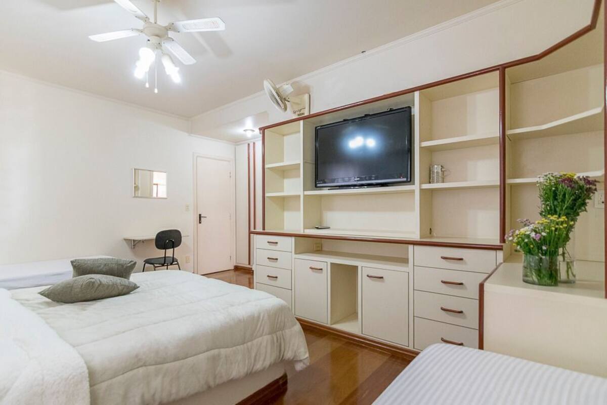 A bedroom with a bed, a desk with a TV, a chair, and a small table with a vase.