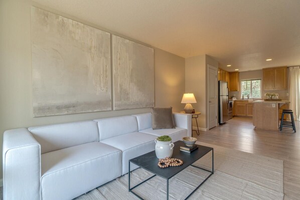 Ein modernes Wohnzimmer mit einem weißen Sofa, einem Couchtisch mit Dekoration und einer Küche im Hintergrund.