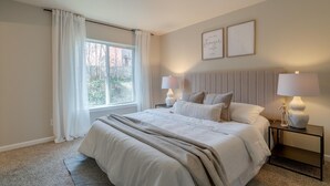Un dormitorio con una cama grande, dos mesitas de noche con lámparas, un ventanal con cortinas y dos citas motivacionales enmarcadas en la pared.